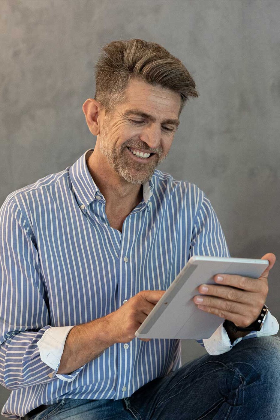 Christos Karakostas in a striped shirt using a tablet, photographed in a minimal studio setting with a neutral gray background.