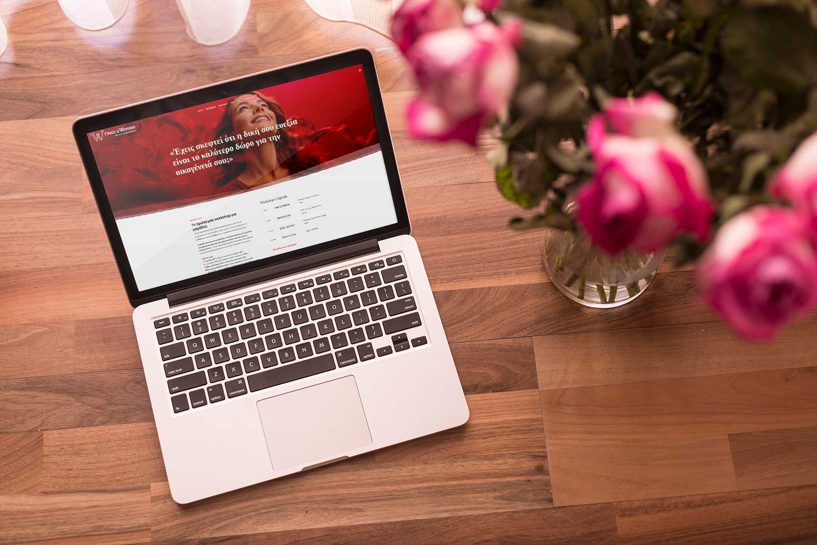 Once a Woman website mockup displayed on a laptop, showing the homepage with a warm red hero section on a wooden desk next to flowers